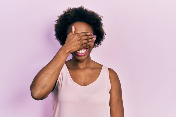 Mladá Afroameričanka v neformální tričku bez rukávů se usmívala a smála se s rukou na tváři, aby zakryla oči překvapením. slepý koncept.  - Fotografie, Obrázek