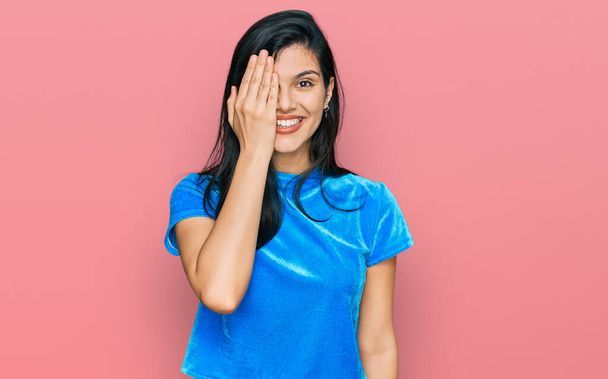 Jeune femme hispanique portant des vêtements décontractés couvrant un oeil avec la main, sourire confiant sur le visage et l'émotion surprise.  - Photo, image