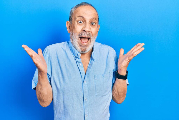 Knappe senior man met baard in casual blauw shirt viert overwinning met vrolijke glimlach en winnaarsuitdrukking met opgeheven handen  - Foto, afbeelding