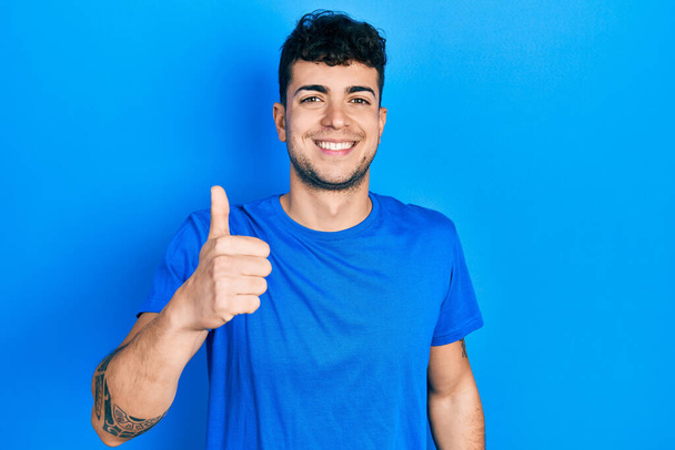 若いヒスパニック系の男性が手で幸せな親指アップジェスチャーを行うカジュアルな青のTシャツを着ています。成功を示すカメラを見ると.  - 写真・画像