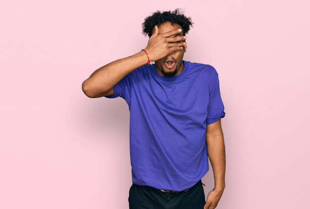 Fiatal afro-amerikai férfi szakállas alkalmi lila pólóban leselkedik sokk borító arc és szemek kézzel, néz keresztül ujjain zavarba ejtő kifejezés.  - Fotó, kép