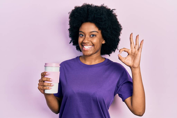 Νεαρή Αφροαμερικανή γυναίκα κρατώντας καφέ κάνει ok υπογράψει με τα δάχτυλα, χαμογελώντας φιλικό gesturing εξαιρετικό σύμβολο  - Φωτογραφία, εικόνα