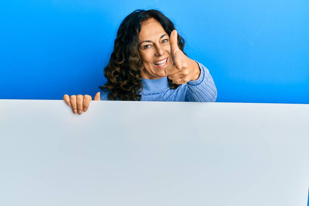 Keski-ikä latino nainen pitämällä tyhjä banneri osoittaa sormella kameraan onnellinen ja hauska kasvot. hyvä energia ja vibes.  - Valokuva, kuva