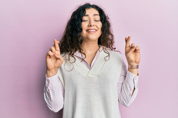Jonge brunette vrouw met krullend haar draagt casual kleding gebaren vinger gekruist glimlachen met hoop en ogen dicht. geluk en bijgelovig concept.  - Foto, afbeelding