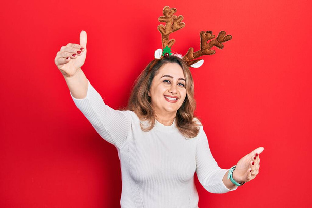 Middle age caucasian woman wearing cute christmas reindeer horns looking at the camera smiling with open arms for hug. cheerful expression embracing happiness.  - Photo, Image