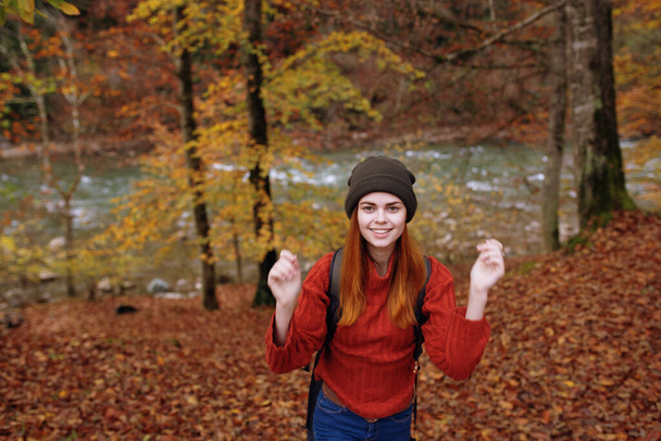 joyful woman in a hat sweater with a backpack on her back gestures with her hands in a park in nature - Photo, image