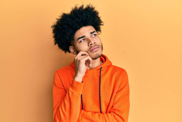 Young african american man with afro hair wearing casual sweatshirt thinking concentrated about doubt with finger on chin and looking up wondering  - Fotoğraf, Görsel
