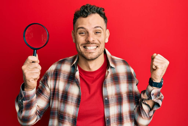 Young hispanic man holding magnifying glass looking for stain at clothes screaming proud, celebrating victory and success very excited with raised arm  - Foto, immagini