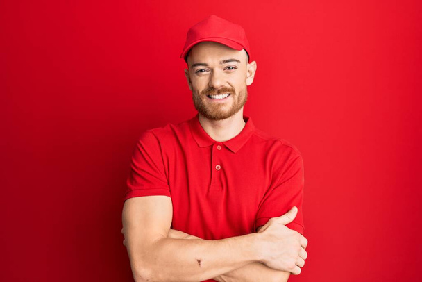 Young redhead man wearing delivery uniform and cap happy face smiling with crossed arms looking at the camera. positive person.  - Foto, Imagen