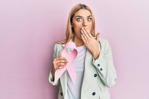 Mujer caucásica joven que usa ropa de negocios con cinta de cáncer rosa que cubre la boca con la mano, conmocionada y temerosa de equivocarse. expresión sorprendida  - Foto, Imagen