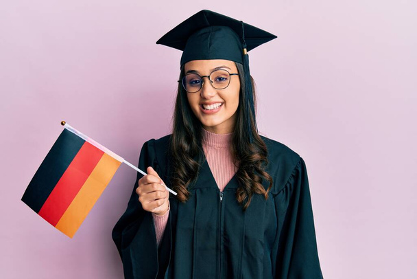 Mujer hispana joven vistiendo uniforme de graduación sosteniendo bandera de Alemania con aspecto positivo y feliz de pie y sonriendo con una sonrisa confiada mostrando los dientes  - Foto, Imagen