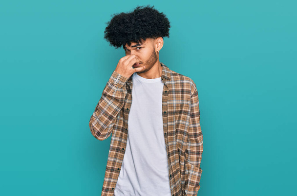 Young african american man with afro hair wearing casual clothes smelling something stinky and disgusting, intolerable smell, holding breath with fingers on nose. bad smell  - Fotó, kép