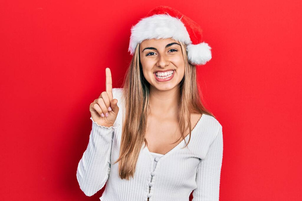 Piękna Latynoska kobieta w świątecznym kapeluszu pokazuje i wskazuje palcem numer jeden uśmiechając się z uśmiechem pewny siebie i szczęśliwy.  - Zdjęcie, obraz