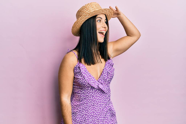 Young brunette woman wearing summer dress and hat very happy and smiling looking far away with hand over head. searching concept.  - Фото, зображення