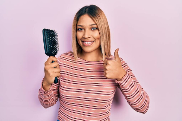 Mulher hispânica bonita segurando escova de cabelo sorrindo feliz e positivo, polegar para cima fazendo excelente e sinal de aprovação  - Foto, Imagem