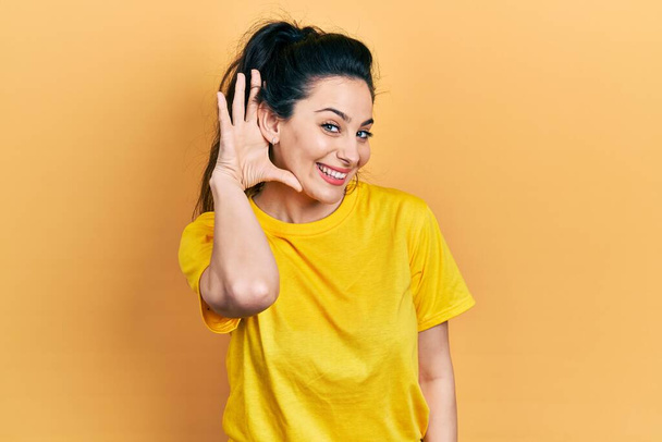 Junge hispanische Frau in lässiger Kleidung lächelt mit der Hand über dem Ohr und hört Gerüchten oder Klatsch zu. Konzept für Gehörlosigkeit.  - Foto, Bild