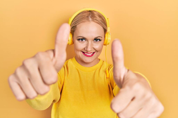 Young blonde woman listening to music using headphones approving doing positive gesture with hand, thumbs up smiling and happy for success. winner gesture.  - 写真・画像