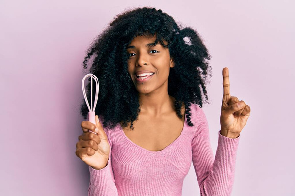 African american woman with afro hair holding small whisk smiling with an idea or question pointing finger with happy face, number one  - Fotó, kép