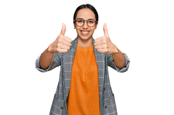 Jong Latijns-Amerikaans meisje draagt een zakelijke jas en een bril die positief gebaar maakt met de hand, glimlacht en blij is voor succes. winnaar gebaar.  - Foto, afbeelding