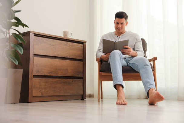 Hombre con libro sentado en sillón en casa. Calefacción por suelo radiante - Foto, imagen
