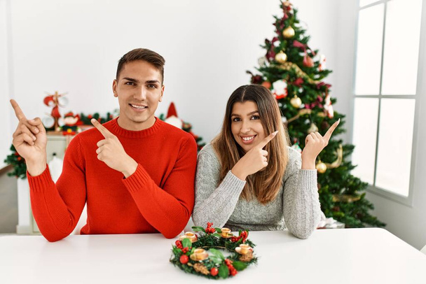 Fiatal spanyol pár ül az asztalnál karácsonykor mosolyog, és a kamerába néz, két kézzel és ujjal oldalra mutatva..  - Fotó, kép