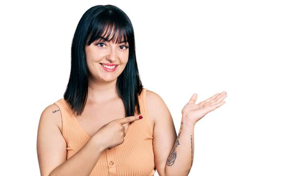 jonge latino plus size vrouw dragen casual kleding verbaasd en glimlachen naar de camera terwijl presenteren met de hand en wijzen met de vinger.  - Foto, afbeelding
