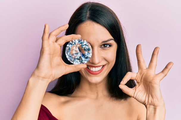 Mulher hispânica jovem segurando pedra de diamante brilhante no olho fazendo sinal ok com os dedos, sorrindo amigável gesto excelente símbolo  - Foto, Imagem