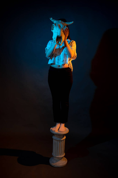 A girl stands on a Greek column with a bull skull in her hands illuminated by blue and yellow light - Foto, immagini