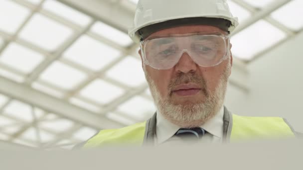 Close-up of male Caucasian foreman with silver beard wearing green reflective vest and hard hat, looking at paper with draft, that reflecting in his protective goggles - Footage, Video