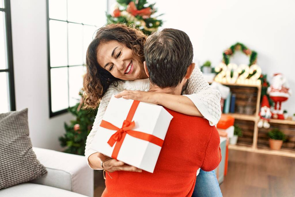 Spanisches Paar mittleren Alters lächelt glücklich und hält Weihnachtsgeschenk zu Hause. - Foto, Bild