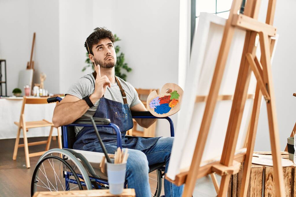 Junger hispanischer Mann, der im Rollstuhl im Kunstatelier sitzt und traurig und aufgebracht nach oben zeigt, mit den Fingern die Richtung anzeigt, unglücklich und deprimiert.  - Foto, Bild
