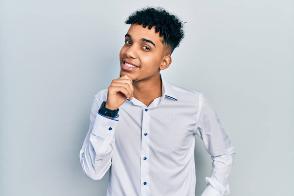 Giovane uomo afroamericano che indossa camicia da lavoro sorridente guardando fiducioso alla fotocamera con le braccia incrociate e mano sul mento. pensare positivo.  - Foto, immagini