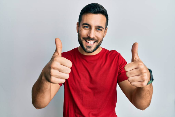 Joven hombre hispano usando ropa casual aprobando hacer gesto positivo con la mano, pulgares arriba sonriendo y feliz por el éxito. gesto ganador.  - Foto, imagen