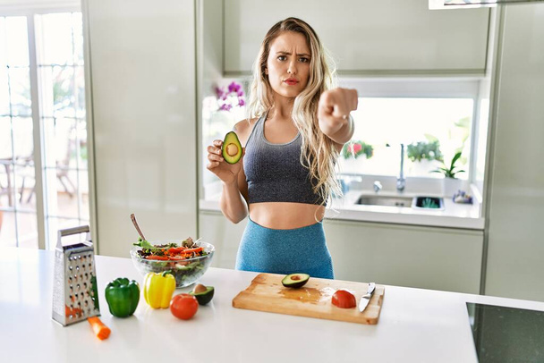 Молодая кавказская фитнес-женщина в спортивной одежде готовит здоровый салат на кухне, указывая пальцем на камеру и на вас, уверенный жест выглядит серьезным  - Фото, изображение