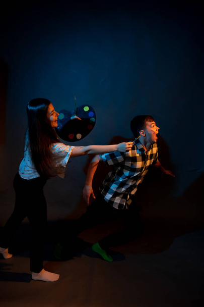 A girl paints a guy in the studio on a dark background illuminated by multicolored light - Photo, image