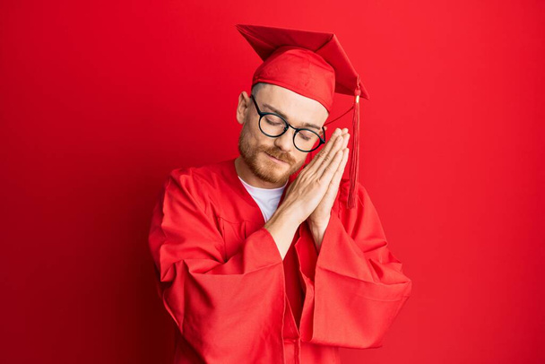 Νεαρός κοκκινομάλλης που φοράει κόκκινο καπέλο αποφοίτησης και επίσημη ρόμπα κοιμάται κουρασμένος ονειρεύεται και ποζάρει με τα χέρια μαζί, ενώ χαμογελά με κλειστά μάτια.  - Φωτογραφία, εικόνα
