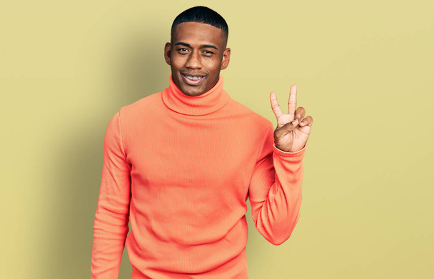 Молодой черный мужчина в оранжевом свитере в водолазке улыбается со счастливым лицом, подмигивая в камеру, делая знак победы. Номер два.  - Фото, изображение