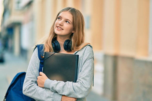 Beautiful caucasian student teenager using headphones holding binder at the city. - Фото, изображение