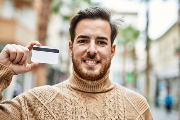 Νεαρός καυκάσιος άνδρας χαμογελά χαρούμενος κρατώντας πιστωτική κάρτα στην πόλη. - Φωτογραφία, εικόνα