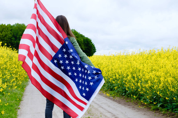 Девушка повернула назад, держа высокий американский флаг на полевой дороге среди жёлтых цветов. Высокое качество фото - Фото, изображение