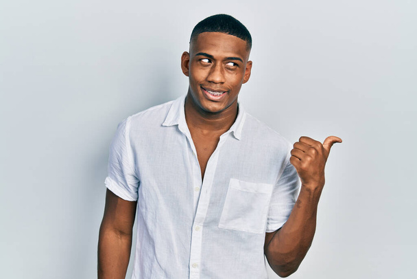 Jeune homme noir portant une chemise blanche décontractée souriant avec le visage heureux regardant et pointant vers le côté avec le pouce vers le haut.  - Photo, image