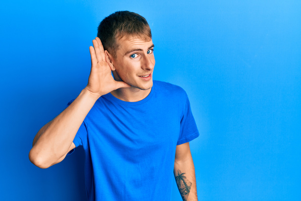 Junger kaukasischer Mann in lässigem blauem T-Shirt, der lächelnd mit der Hand über dem Ohr Gerüchten oder Klatsch lauscht. Konzept für Gehörlosigkeit.  - Foto, Bild
