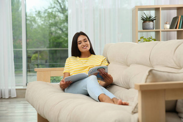 Young woman reading magazine on sofa at home - Foto, imagen