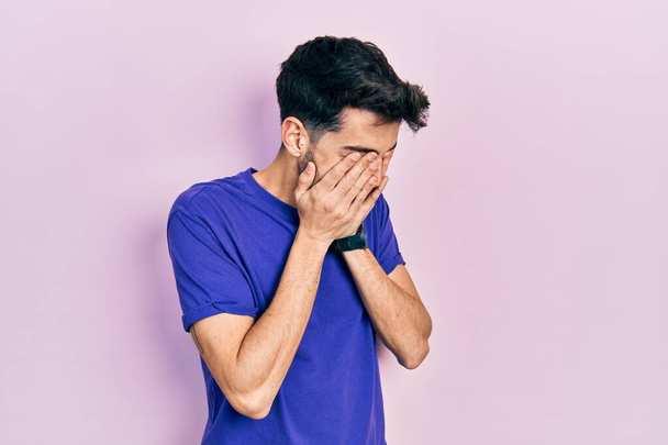 Junger hispanischer Mann in lässigem T-Shirt mit traurigem Gesichtsausdruck, der das Gesicht mit Händen bedeckt, während er weint. Depressionskonzept.  - Foto, Bild
