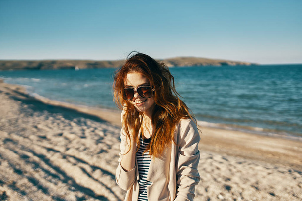 海の近くの眼鏡をかけたビーチの赤い髪の女性ベージュコートTシャツモデル - 写真・画像