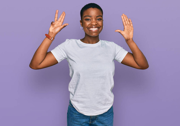 Молода африканка в білій сорочці з білою сорочкою показує і вказує вгору пальцями номер дев'ять, посміхаючись впевнено і щасливо.  - Фото, зображення