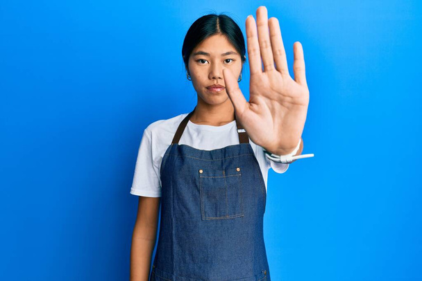 Giovane donna cinese che indossa grembiule cameriere facendo smettere di cantare con il palmo della mano. espressione ammonitrice con gesto negativo e serio sul viso.  - Foto, immagini