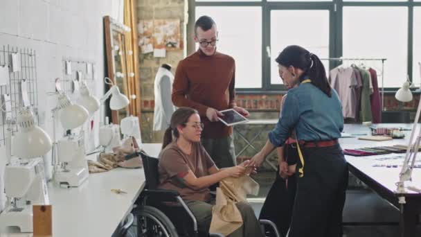 Slowmo médio tiro de costureira feminina alegre em cadeira de rodas sorrindo e mostrando vestuário para a equipe de designers de moda em estúdio - Filmagem, Vídeo