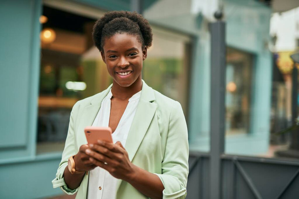 Joven mujer de negocios afroamericana sonriendo feliz usando el teléfono inteligente en la ciudad. - Foto, imagen