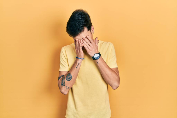 Jovem hispânico vestindo casual camiseta amarela esfregando os olhos para fadiga e dor de cabeça, sonolento e expressão cansada. problema de visão  - Foto, Imagem